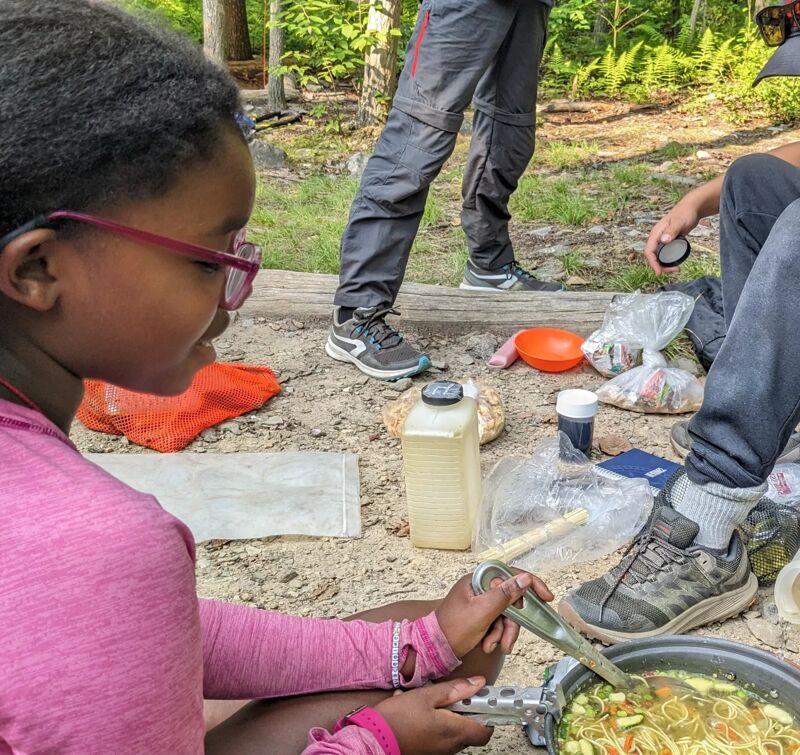 A young girl with glasses and a pink shirt is stirring a pot of soup while camping. Other people are in the background, along with camping gear like a water container, bowls, and bags of food. The setting appears to be a forest, with trees visible in the background.